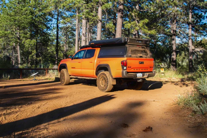 An orange Toyota Tacoma overland vehicle, functioning as an RV rental, parked amidst tall pines for camping.. Toyota Tacoma TRD Pro 2023