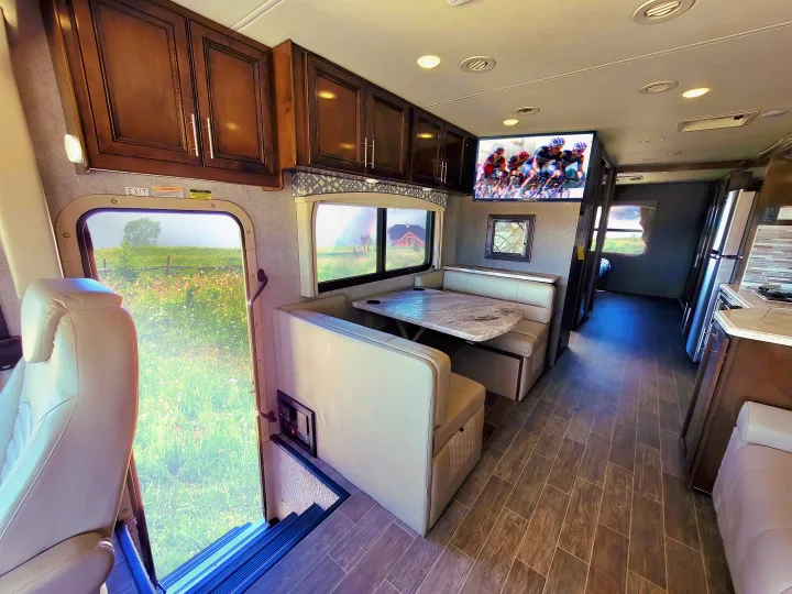 Inside a modern RV rental, featuring a full dinette with a TV, rich wood cabinets, and sleek flooring. Scenic views visible through windows.. Thor Motor Coach Miramar 2020