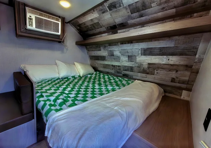 Cozy RV rental bedroom featuring a comfortable bed with a green and white comforter, wall-mounted AC, and rustic wood paneling.. Coleman Lantern 2023