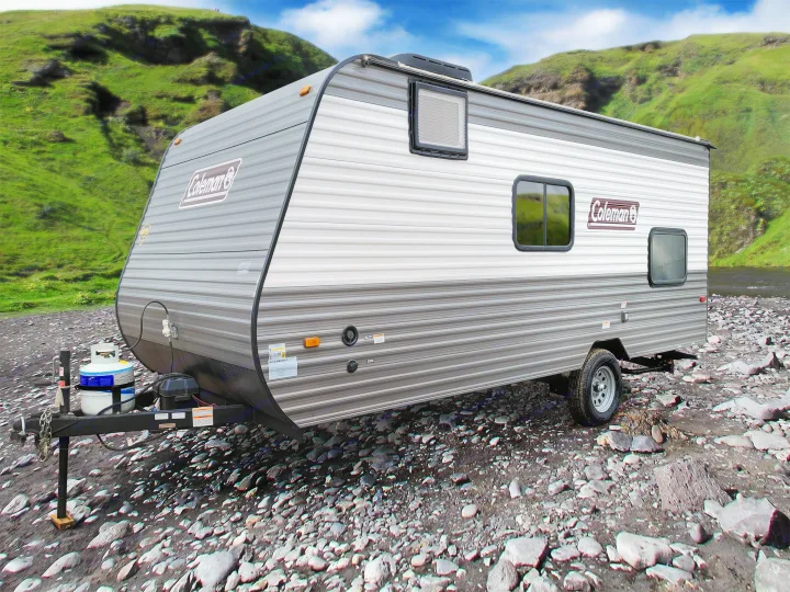 A Coleman travel trailer RV rental parked on a rocky riverbank, surrounded by lush green hills. Ideal for outdoor adventures.. Coleman Lantern 2023