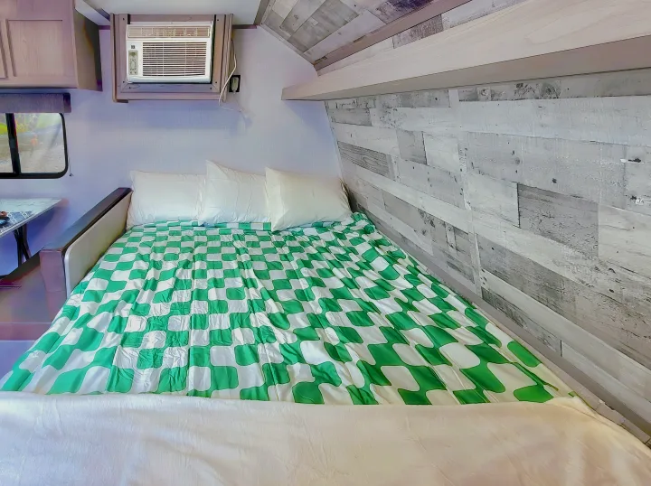 Cozy RV rental interior featuring a bed with green checker comforter, window view, and AC unit.. Coleman Lantern 2023