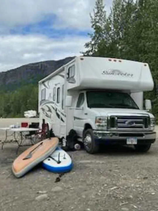 This RV rental is ready for adventure! A white camper with paddleboards and a bike is set up at a scenic campsite.. Forest River Sunseeker 2012
