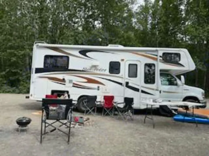 This white RV rental is set up at a serene campsite with chairs, a table, and grill for outdoor relaxation and travel.. Forest River Sunseeker 2012