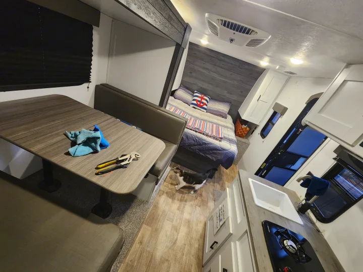 Inside an rv rental: functional dinette with tools, a comfy bed with nautical decor, kitchenette, & AC. A cat explores the interior.. Keystone RV Hideout 2021