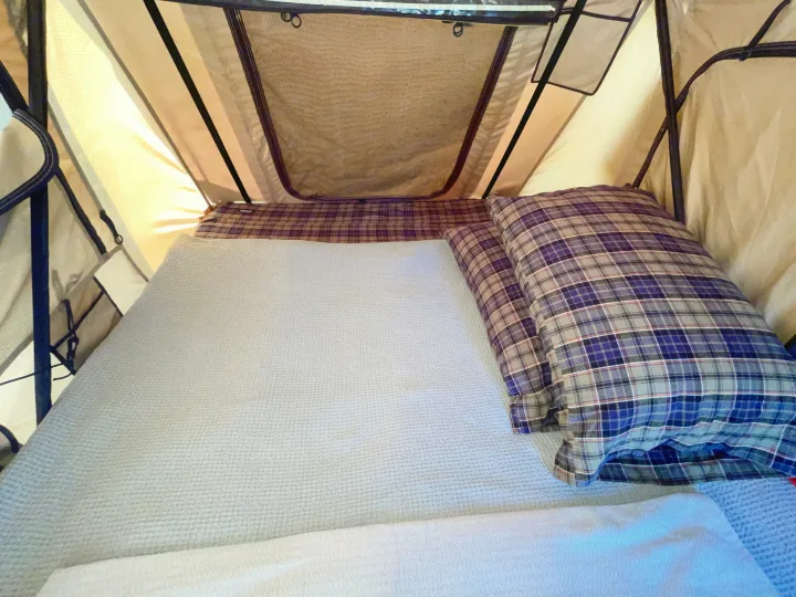 RV rental sleeping space: A cozy bed with a light grey blanket and blue plaid pillows inside a well-lit rooftop tent.. Jeep Patriot 2016