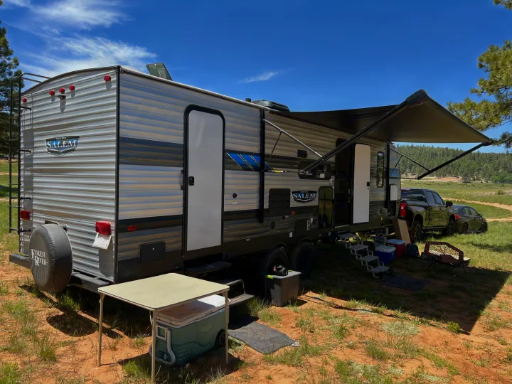 A Forest River Salem RV rental is set up with an extended awning, alongside a truck & car, at a sunny outdoor campsite.. Forest River Salem 2022