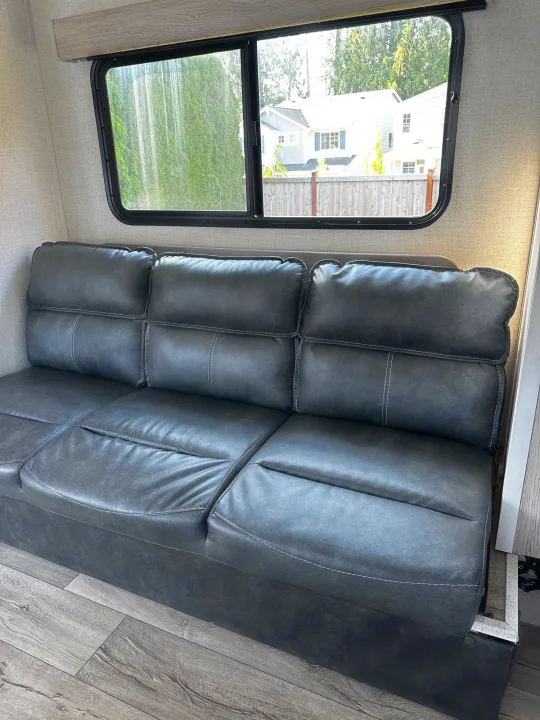 Cozy gray sofa inside an RV rental, offering comfortable seating with a view of trees and houses through the window.. Dutchmen Coleman Rubicon 2021