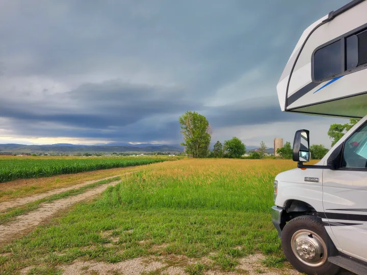 An RV rental overlooks vibrant green fields and distant mountains under a dramatic cloudy sky from a rural dirt path.. Solera Solera Motorhome 2022