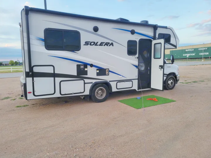 White Solera RV rental parked on gravel near an exposition center, door open, with a green mat outside. Ready for adventure!. Solera Solera Motorhome 2022