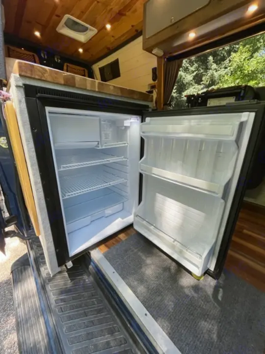 Inside this RV rental: open compact refrigerator showing freezer and fridge shelves. Perfect camper van kitchen setup.. Mercedes Sprinter 2016
