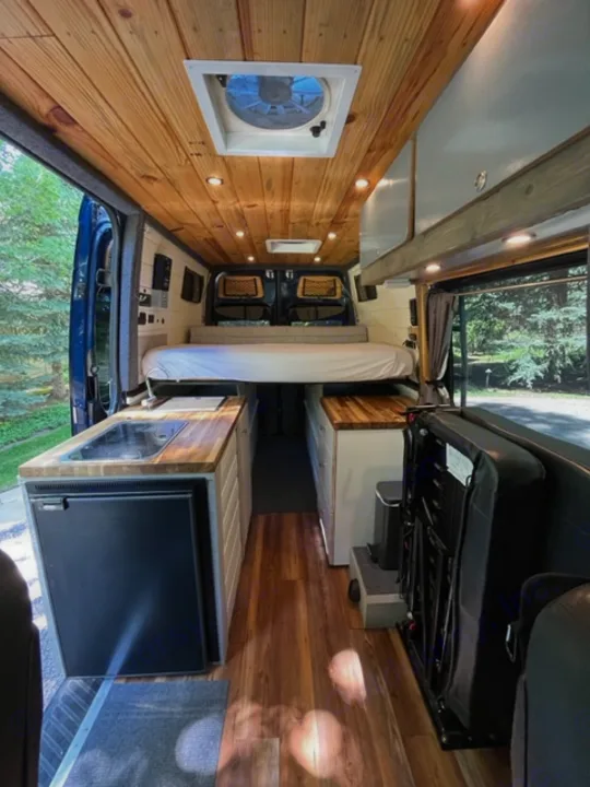 Explore this bright camper van RV rental interior! Features wood walls, a fan, kitchen with fridge, and flexible seating/bed area.. Mercedes Sprinter 2016