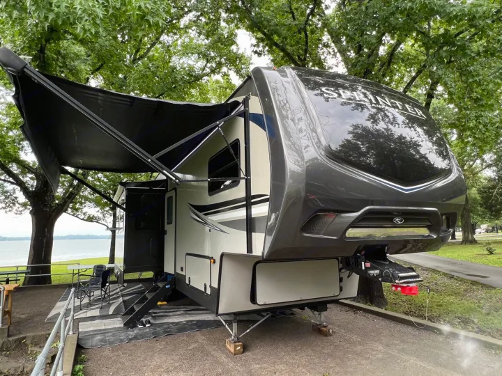 A grey Sprinter fifth wheel RV rental parked by a lake, with an extended awning and outdoor chairs for lakeside camping.. Keystone RV Sprinter Campfire Edition 2020