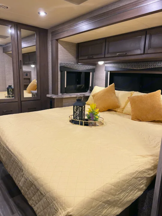 Relax in this cozy **rv rental** bedroom. Features a comfortable bed with a light yellow quilt, mirrored wardrobe, and dark wood cabinets.. Thor Four Winds 2021