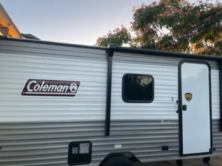 A modern Coleman travel trailer, white and grey, with a side door and window. Explore the outdoors with this RV rental option.. Dutchmen Coleman Lantern LT 2023