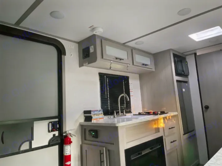 Compact RV rental kitchen features a sink, stove, microwave, and fridge. Fire extinguisher visible near the entry door.. Dutchmen Coleman Lantern LT 2023