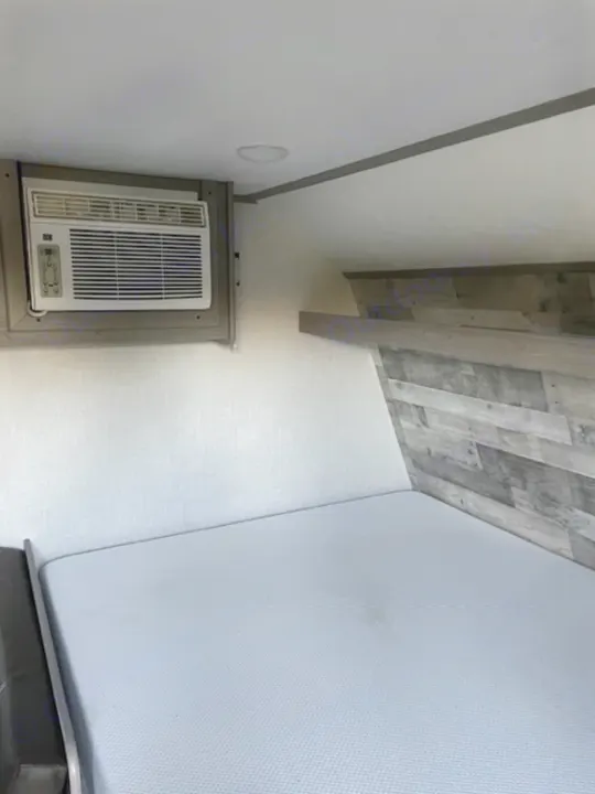 Inside a cozy RV rental: compact sleeping area features a fresh mattress, wall-mounted AC unit, and stylish wood-look wall.. Dutchmen Coleman Lantern LT 2023