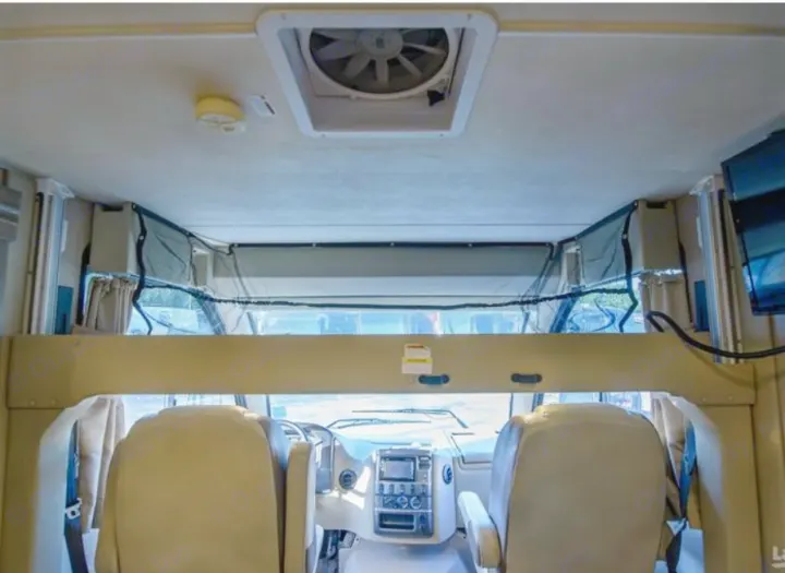 Inside this RV rental: view of the front cockpit from the living area, featuring an overhead bunk with safety net, ceiling fan, and driver's dashboard. A comfortable motorhome.. Thor Vegas 2018