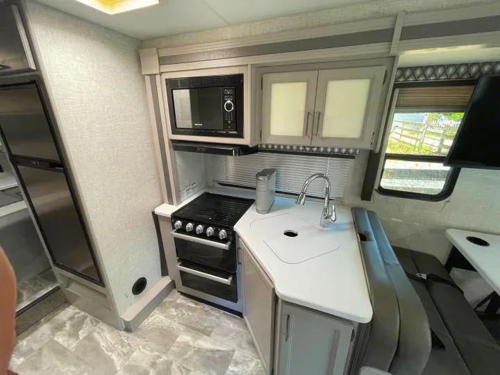 Modern RV rental kitchen and dinette with microwave, stovetop, sink, and light grey seating. Bright, compact interior.. Thor Quantum LC 2022