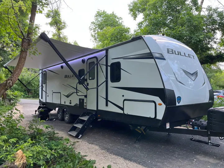 A white Bullet travel trailer RV rental at a green campsite. Its extended awning features purple lights, and steps lead inside.. Keystone RV Bullet Ultra Lite 2022