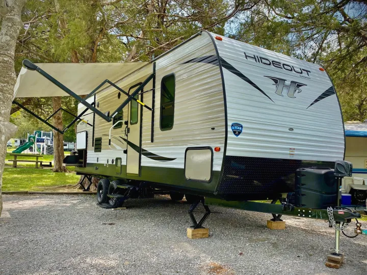 A white Hideout travel trailer RV, with its awning extended, sits on a gravel campsite. Ready for your next RV rental trip.. Keystone RV Hideout LHS 2019