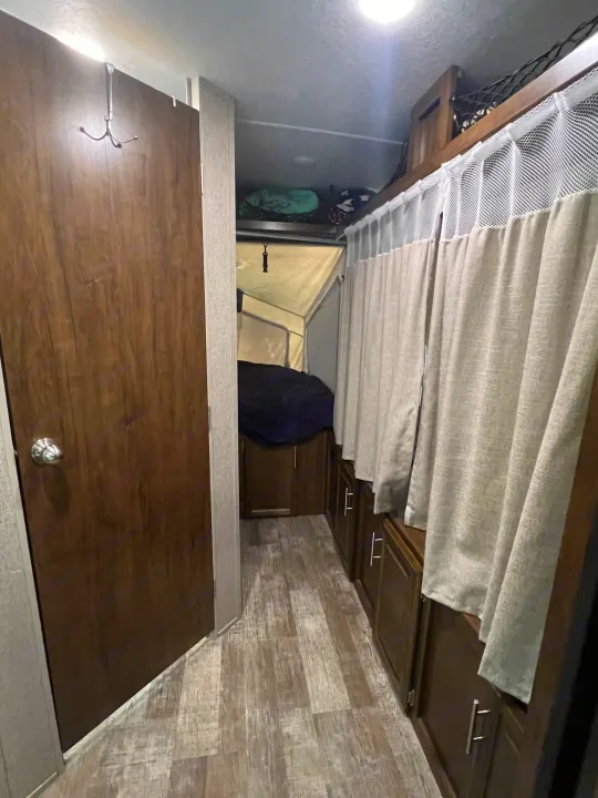 Inside an RV rental, a hallway features a wooden door, laminate flooring, and cabinets with curtains, leading to a pop-up bed area.. Forest River Rockwood Roo 2019