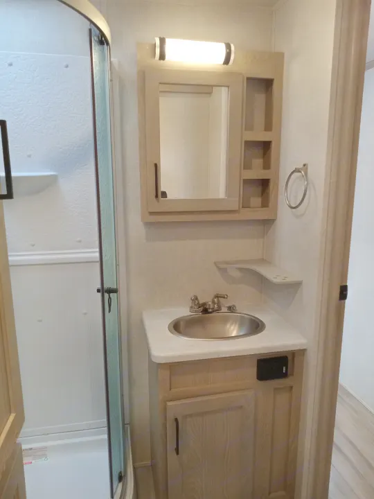 Compact RV rental bathroom featuring a glass-enclosed shower, vanity with stainless steel sink, mirror, and light wood cabinetry.. Forest River Rockwood Signature 2021