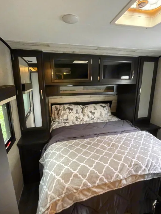RV rental bedroom with a bed, moose-patterned pillows, dark wood cabinets, and overhead vent.. Jayco Jay Feather 2021