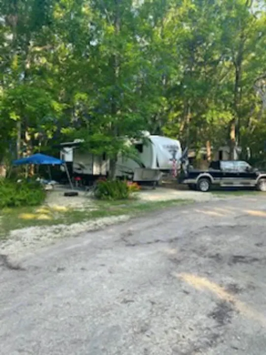 An RV rental, a fifth-wheel camper, is set up with a truck under trees at a shaded campsite, featuring a blue umbrella.. Forest River Sabre 2022
