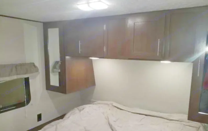 Cozy RV rental bedroom featuring dark wood overhead cabinets, bright lighting, and a comfortable bed, ideal for travel.. Avenger ATI 2018
