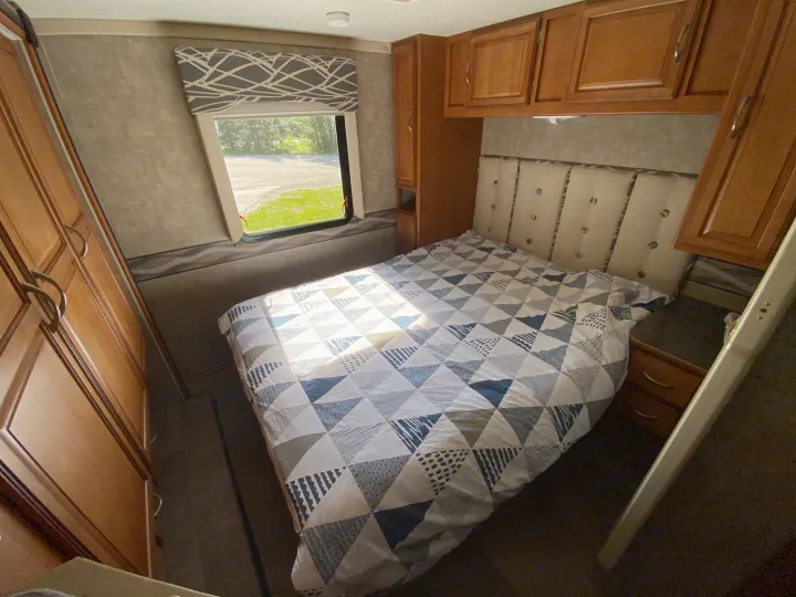 RV rental bedroom with a blue & white patterned bed, wood cabinets, and window looking out to trees & road. Sunlight streams in.. Winnebago Minnie Winnie 2015