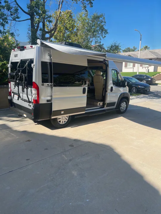 Silver camper van for RV rental, parked in a sunny driveway. Side door open, awning extended, bike rack on rear.. Thor Motor Coach Other 2023