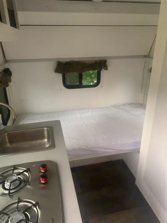 Compact RV rental interior featuring a comfy bed, window with nature views, and a small kitchen with sink and stovetop.. Travel Lite Rove 14BH 2024