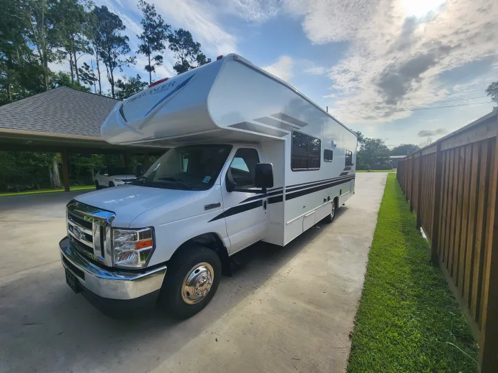Daytime view: White Class C rv rental parked on a concrete driveway, alongside a green lawn and wooden fence.. Coachmen Freelander 2023