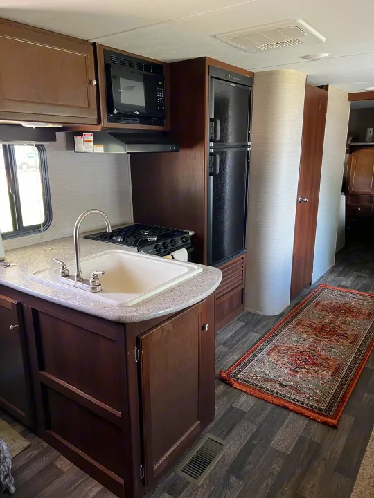 Modern RV rental kitchen: sink, stovetop, microwave, fridge, wood cabinets, and a colorful rug.. Keystone RV Bullet Ultra Lite 2016