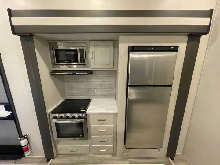 Compact RV rental kitchen featuring a microwave, stove with oven, and full-size refrigerator, with a fire extinguisher nearby.. Keystone Sprinter 2022