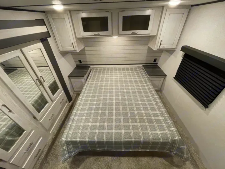 Comfortable RV rental bedroom with a plaid bed, overhead cabinets, nightstands, and a mirrored wardrobe on the left.. Keystone Sprinter 2022