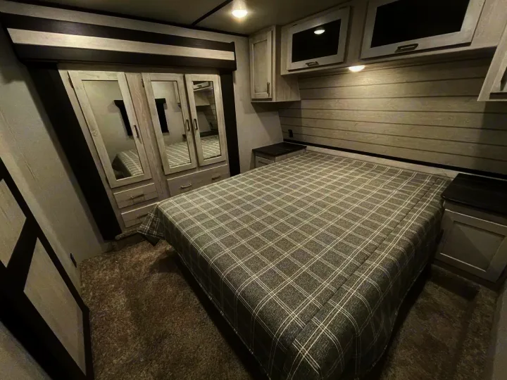 Experience comfort in this modern RV rental bedroom featuring a large plaid bed, mirrored wardrobe, and overhead storage.. Keystone Sprinter 2022