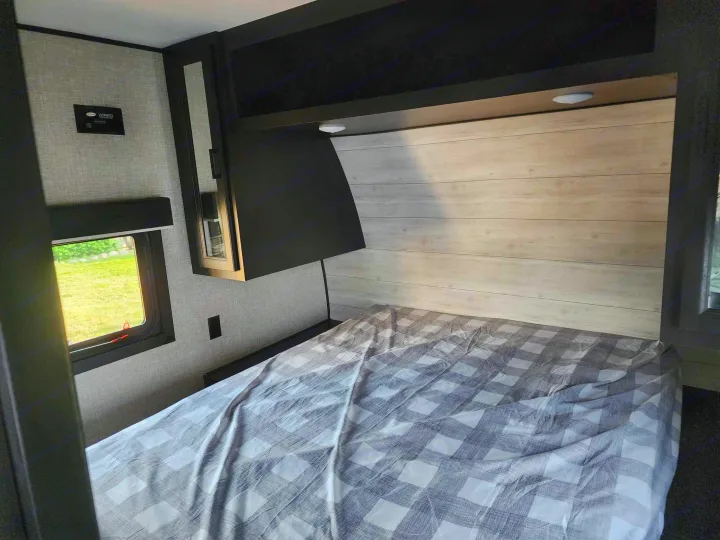 RV rental bedroom featuring a checkered bed, compact overhead storage, and a bright window view.. Jayco Jay Flight 2023