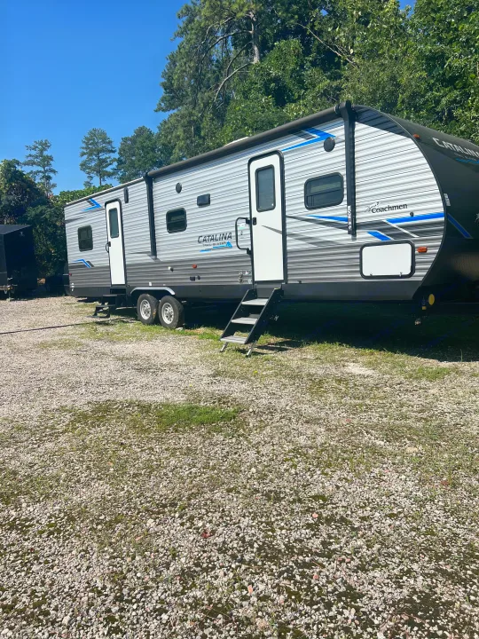 A spacious Coachmen Catalina Trail Blazer RV rental, a silver travel trailer with blue stripes, parked on a gravel lot.. Coachmen Catalina 2021