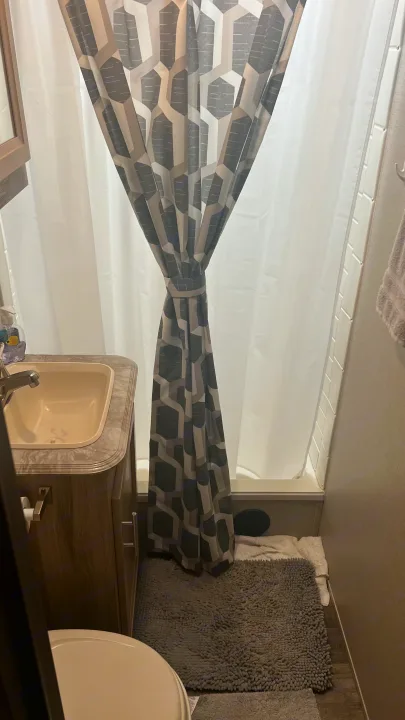 This RV rental bathroom features a sink, toilet, and a geometric-patterned shower curtain tied back, with a gray bathmat.. Coachmen Catalina 2021