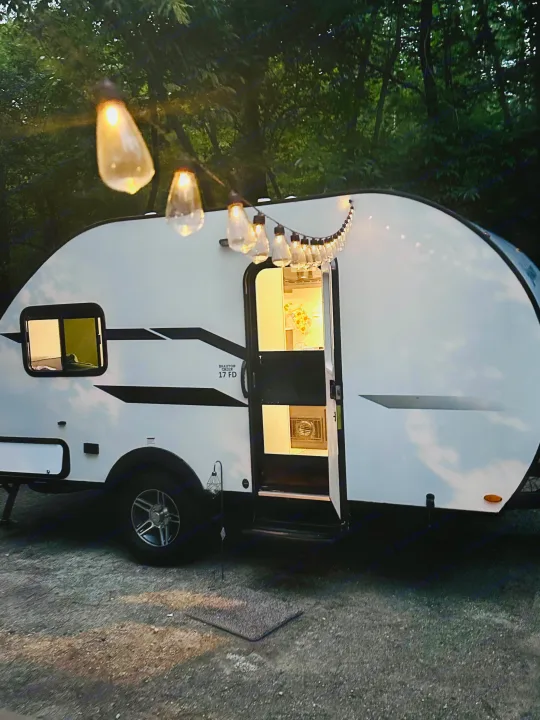 An RV rental glows warmly with string lights at dusk, offering a cozy retreat in a tranquil wooded campsite.. Braxton Creek Bushwhacker Plus 2021