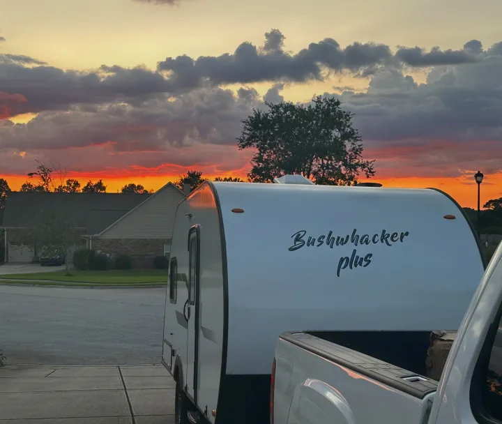 Bushwhacker Plus RV rental hitched to a truck in a driveway. Features a vibrant orange sunset and dramatic clouds.. Braxton Creek Bushwhacker Plus 2021
