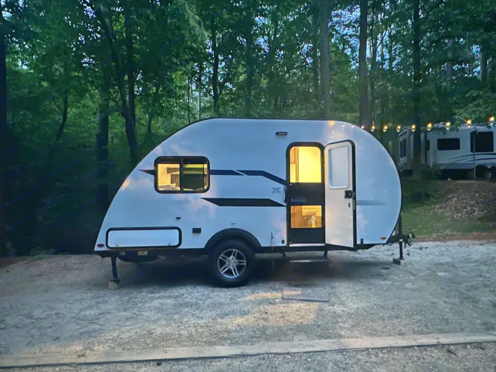 A cozy white teardrop RV rental glows from within, parked at a wooded campsite during the evening.. Braxton Creek Bushwhacker Plus 2021