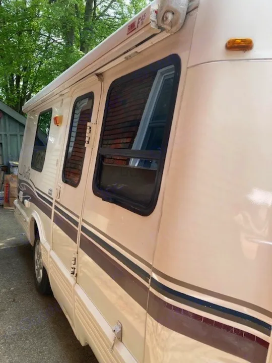 Cream-colored rv rental, side view, with multi-toned stripes, dark windows, door, and a rolled-up awning.. Winnebago Rialta 1998
