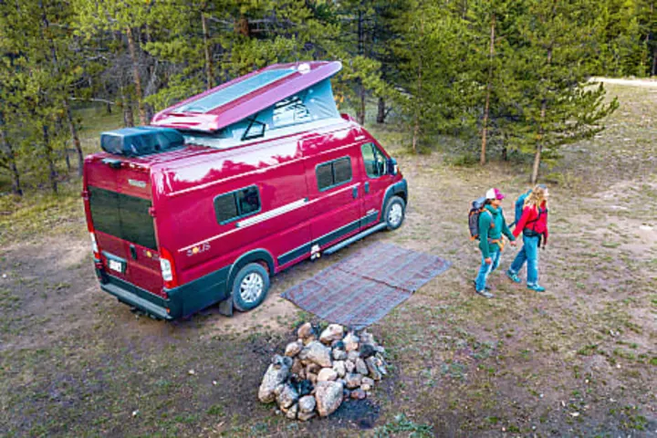 Red RV rental camper van at forest campsite with two people walking and fire pit. Ready for outdoor adventure.. Winnebago Solis PX 2024