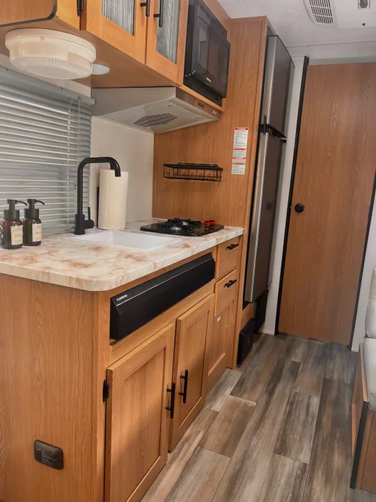Bright RV rental kitchen showcasing light wood cabinets, stainless appliances, a sink, and stylish wood-look flooring.. Avenger LT 2022