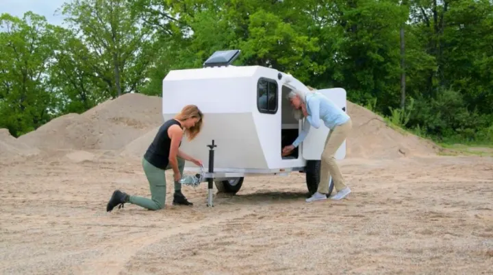 Two women prepare a white teardrop rv rental. One handles the hitch, the other accesses the side storage of the compact camper.. Other Other 2022