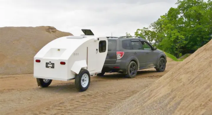 A white teardrop RV rental hitched to a dark SUV on a dirt path with sand piles.. Other Other 2022
