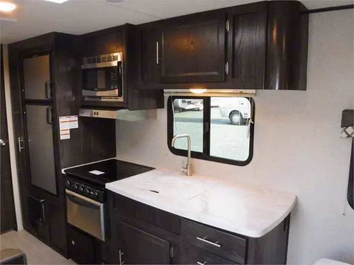 An RV rental kitchen features dark wood cabinets, stainless steel appliances, and a white countertop with a sink.. Dutchmen Kodiak Ultimate 2018