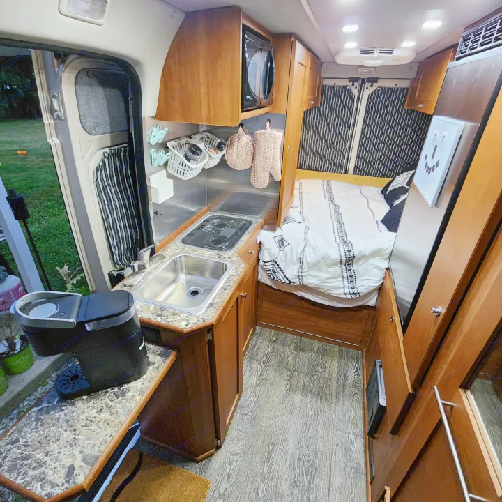 An RV rental's warm interior: right, a bed with patterned bedding; left, a kitchen area with sink, stovetop, and microwave.. Roadtrek Roadtrek Class B 2017